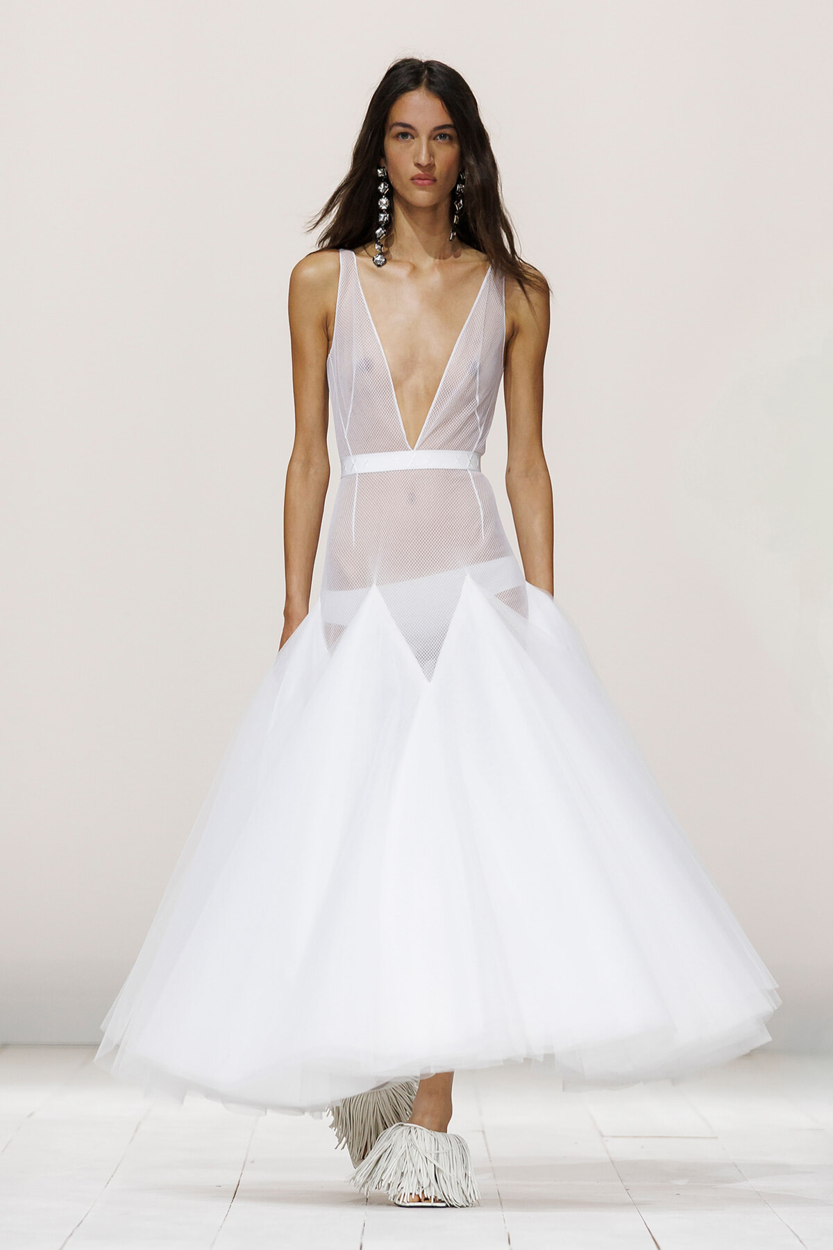 Model wearing a white mesh-plunge gown with a deep V bodice, belt, and voluminous tulle skirt on a runway.