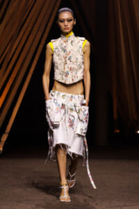 Model walking a fashion runway in a white floral-embroidered sleeveless top and matching distressed skirt with side pockets and colorful piping.