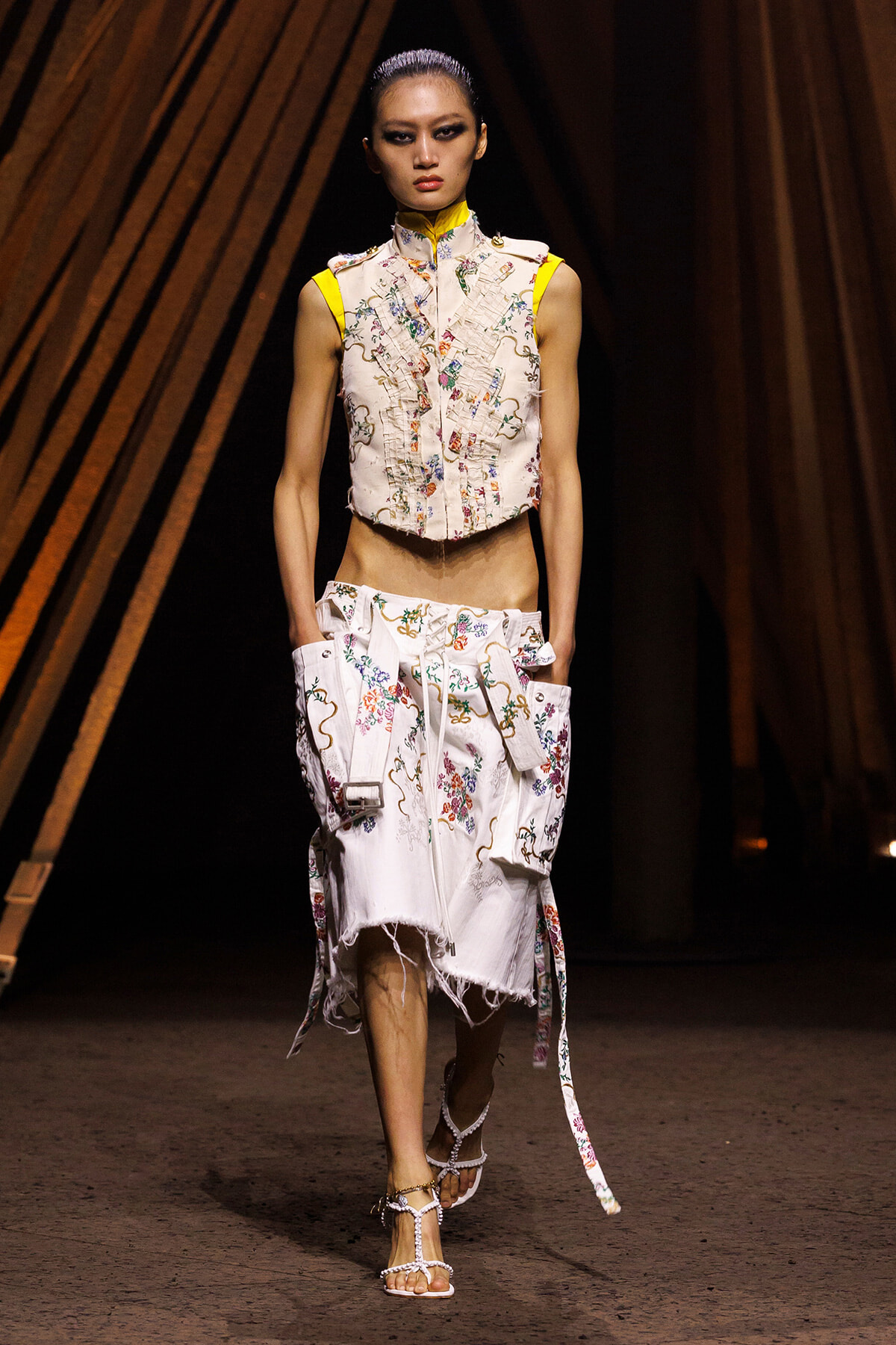 Model walking a fashion runway in a white floral-embroidered sleeveless top and matching distressed skirt with side pockets and colorful piping.
