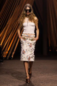 Model walking a fashion runway in a white strapless textured top and embroidered midi skirt with floral motifs, wearing brown open-toe heels and oversized black sunglasses.