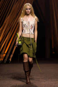 Model on a runway wearing a cream floral corset top, olive cargo skirt, brown knee-high boots, and a neon-green handbag.