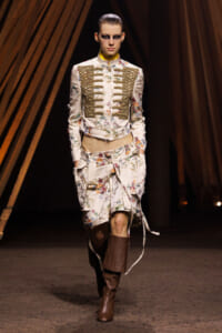 Model on runway wearing a cream floral military-style jacket with gold braid details, matching floral skirt, and brown knee-high boots, under warm stage lighting.