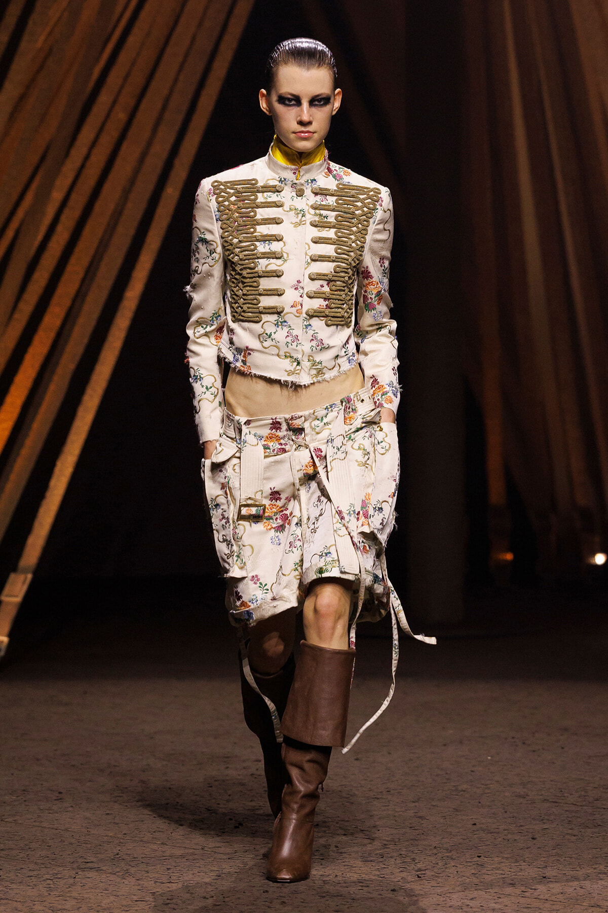 Model on runway wearing a cream floral military-style jacket with gold braid details, matching floral skirt, and brown knee-high boots, under warm stage lighting.