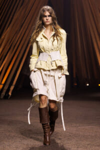 Model walking a runway in a cream utility jacket, white belt, and light shorts with knee-high brown boots.