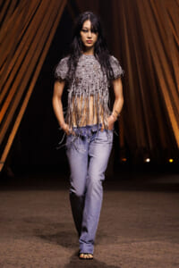 Female model walks the runway in a metallic fringe top paired with loose blue jeans, hands in pockets, dark hair and a confident expression.