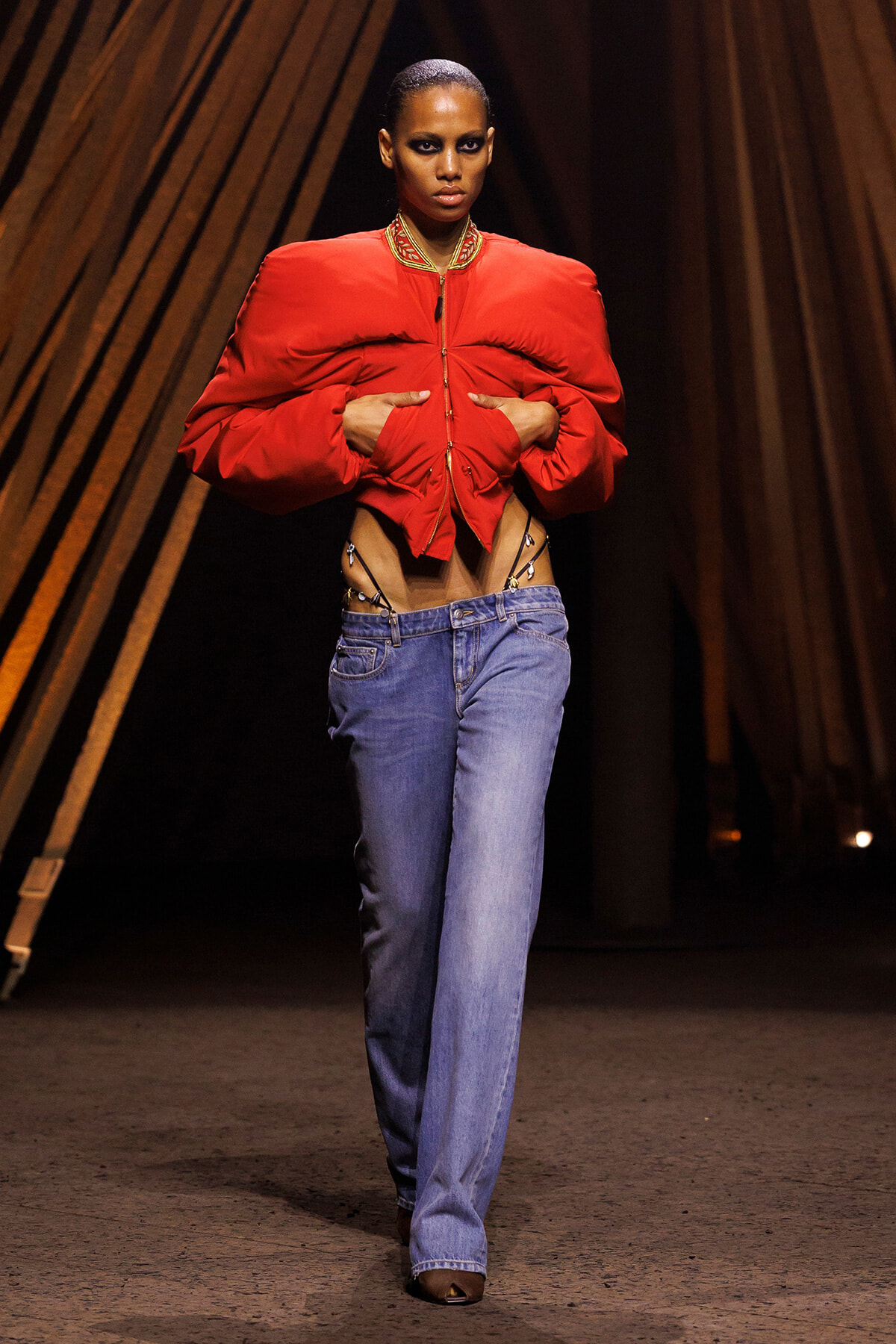 Model on a runway wearing a bold red cropped puffer jacket, midriff exposed, paired with blue jeans. Hands pull the jacket open as she walks.
