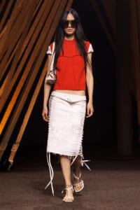 Model on a runway wearing a red sleeveless top, white distressed midi skirt with side lacing, and white sandals with a small shoulder bag and oversized sunglasses.