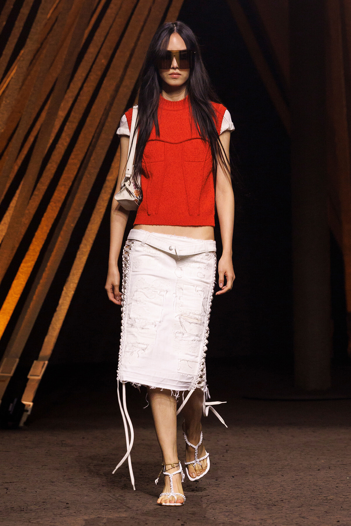 Model on a runway wearing a red sleeveless top, white distressed midi skirt with side lacing, and white sandals with a small shoulder bag and oversized sunglasses.