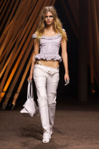 Model wearing a light purple off-shoulder ruffled top and distressed white jeans walks a runway, carrying a white bag.