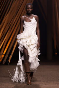 Model walking a runway in a white, ruffled midi dress with an asymmetrical neckline and fringe bag, brown boots visible behind.