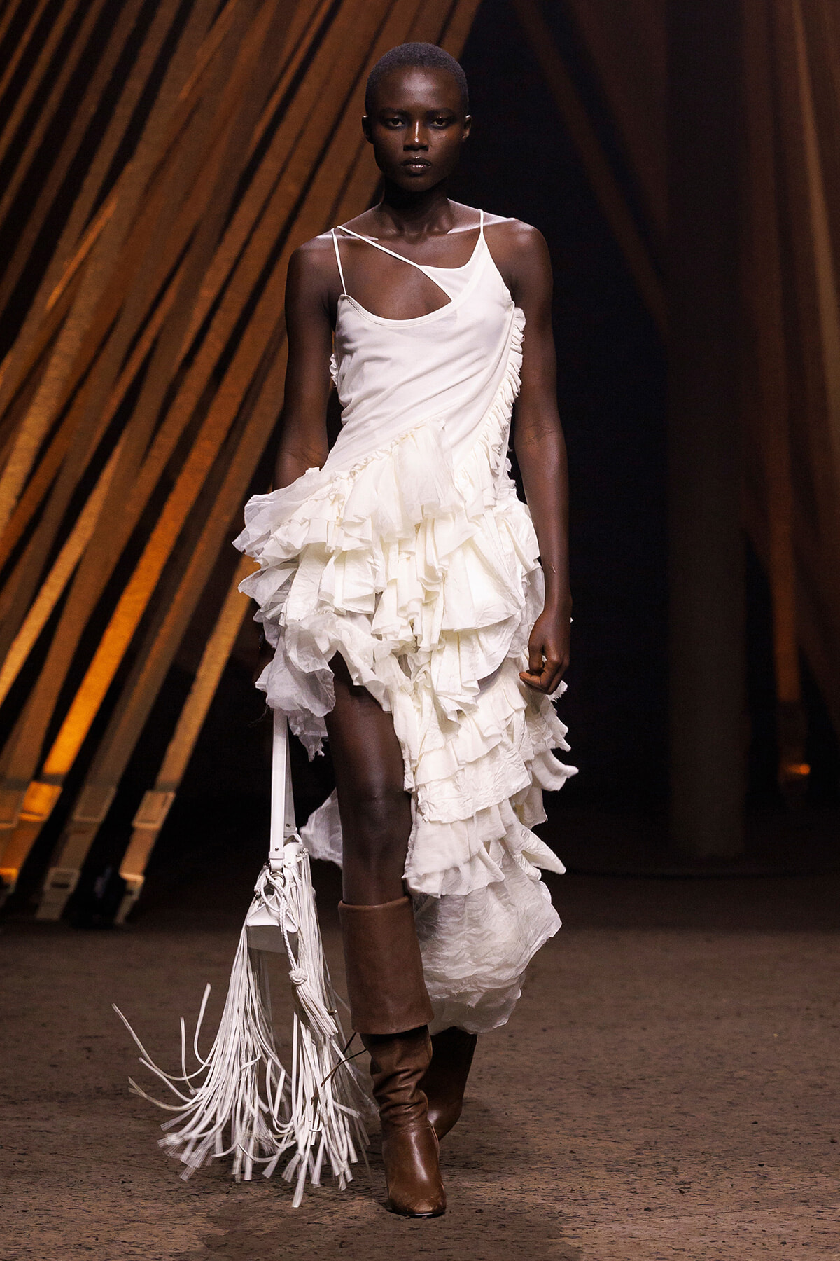 Model walking a runway in a white, ruffled midi dress with an asymmetrical neckline and fringe bag, brown boots visible behind.