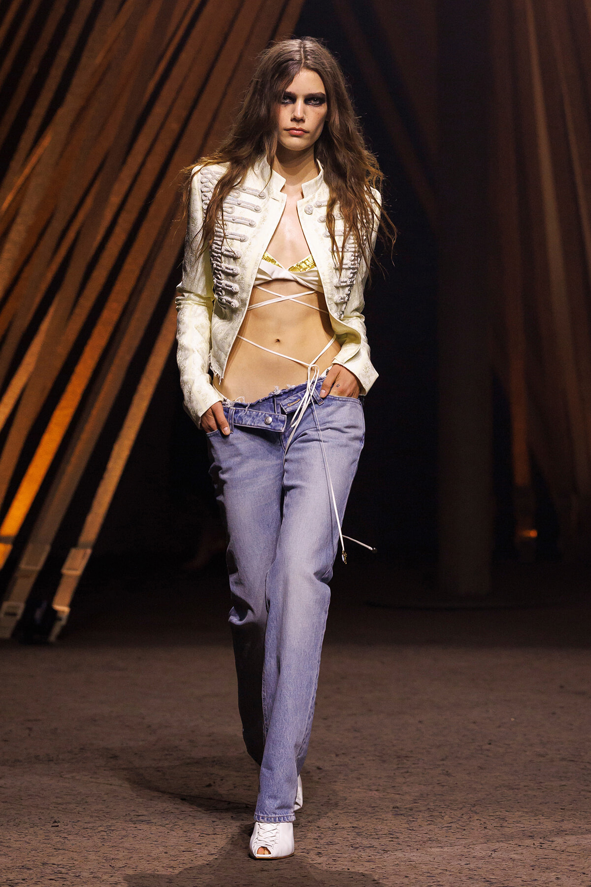 Model walks runway wearing a white military-style cropped jacket over a strappy bikini top and low-rise jeans, white open-toe boots.