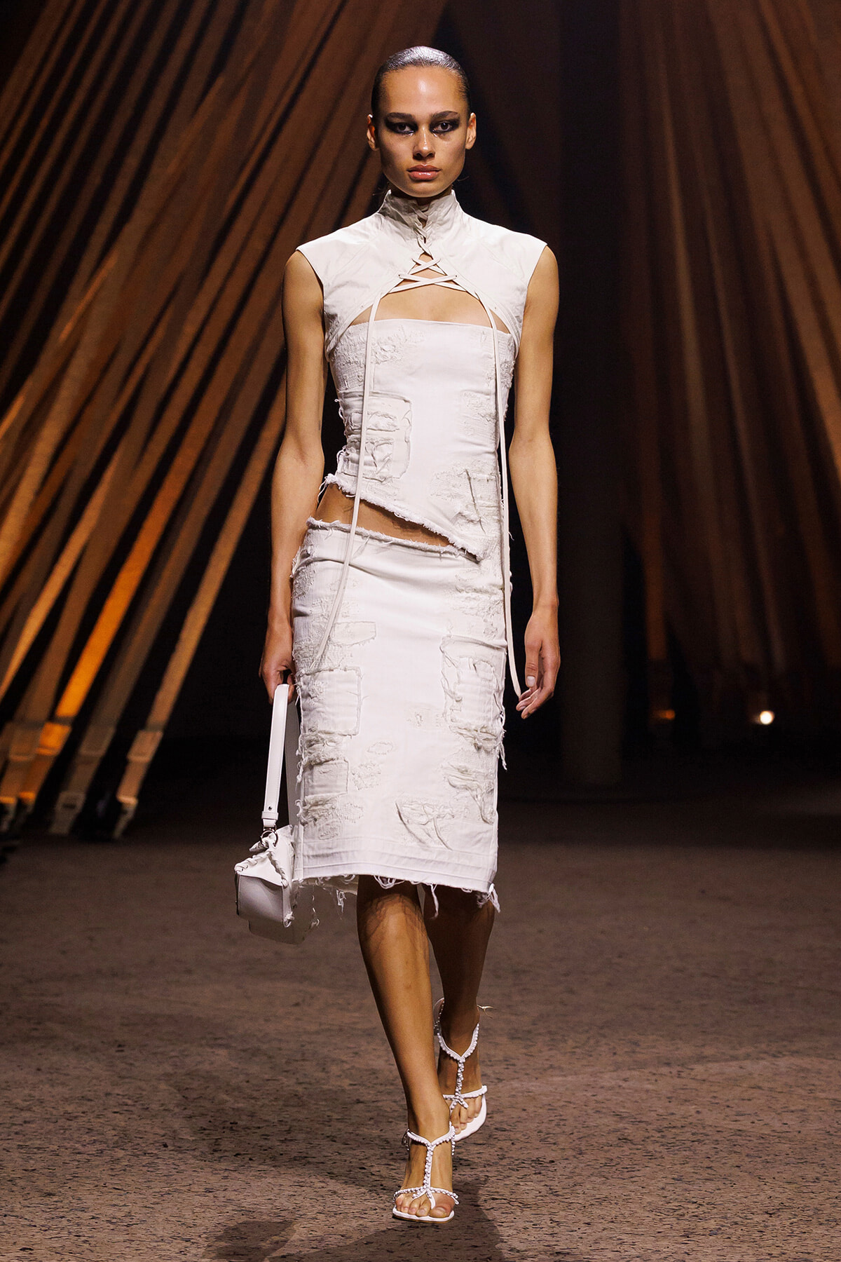 Model on a runway wearing a white distressed two-piece dress with a high neck, crisscross front, and a small white bag, wearing strappy sandals.