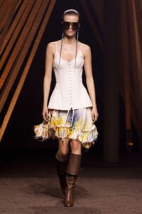 Model on runway wearing a white corset top with lace-up front, a colorful ruffled skirt, knee-high brown boots, and oversized dark sunglasses with a sleek hairdo.