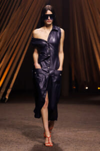 Model walks a runway in a dark navy leather one-shoulder dress with pockets and a front button panel, wearing oversized black sunglasses and orange strappy sandals.