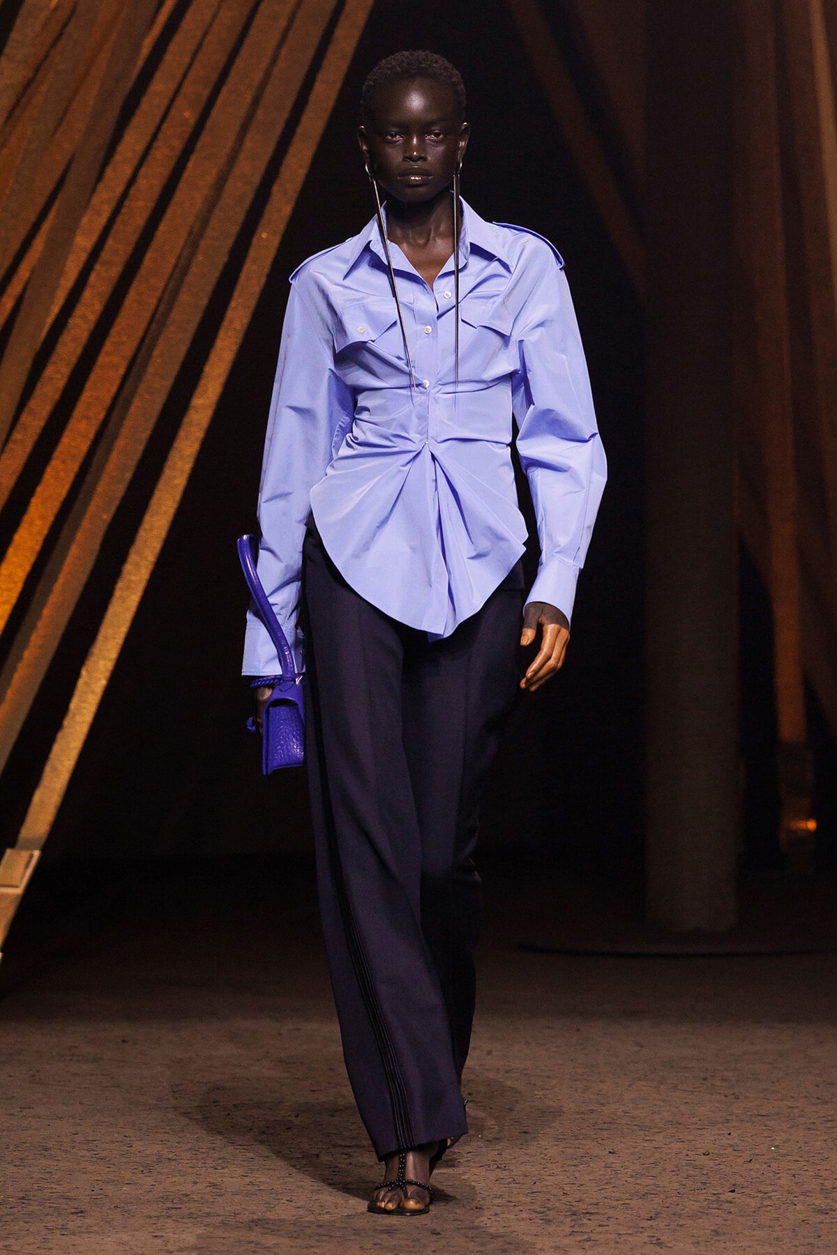 Model walks the runway in a light blue blouse with a draped knot, dark wide-leg pants, and a small purple handbag; minimalist sandals complete the look.