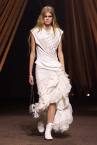 Model walks the runway in a white avant-garde dress with a sculpted bodice, layered ruffles, and white sneakers, carrying a small white tassel purse.