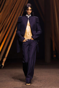 Runway model in a navy military-style jacket with fringe epaulettes and matching pants; jacket open to reveal a bare midriff on a dark catwalk.