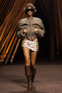 Model struts a runway in a taupe oversized jacket, a white embroidered mini skirt with gold lacing, and tall brown boots, wearing large sunglasses.
