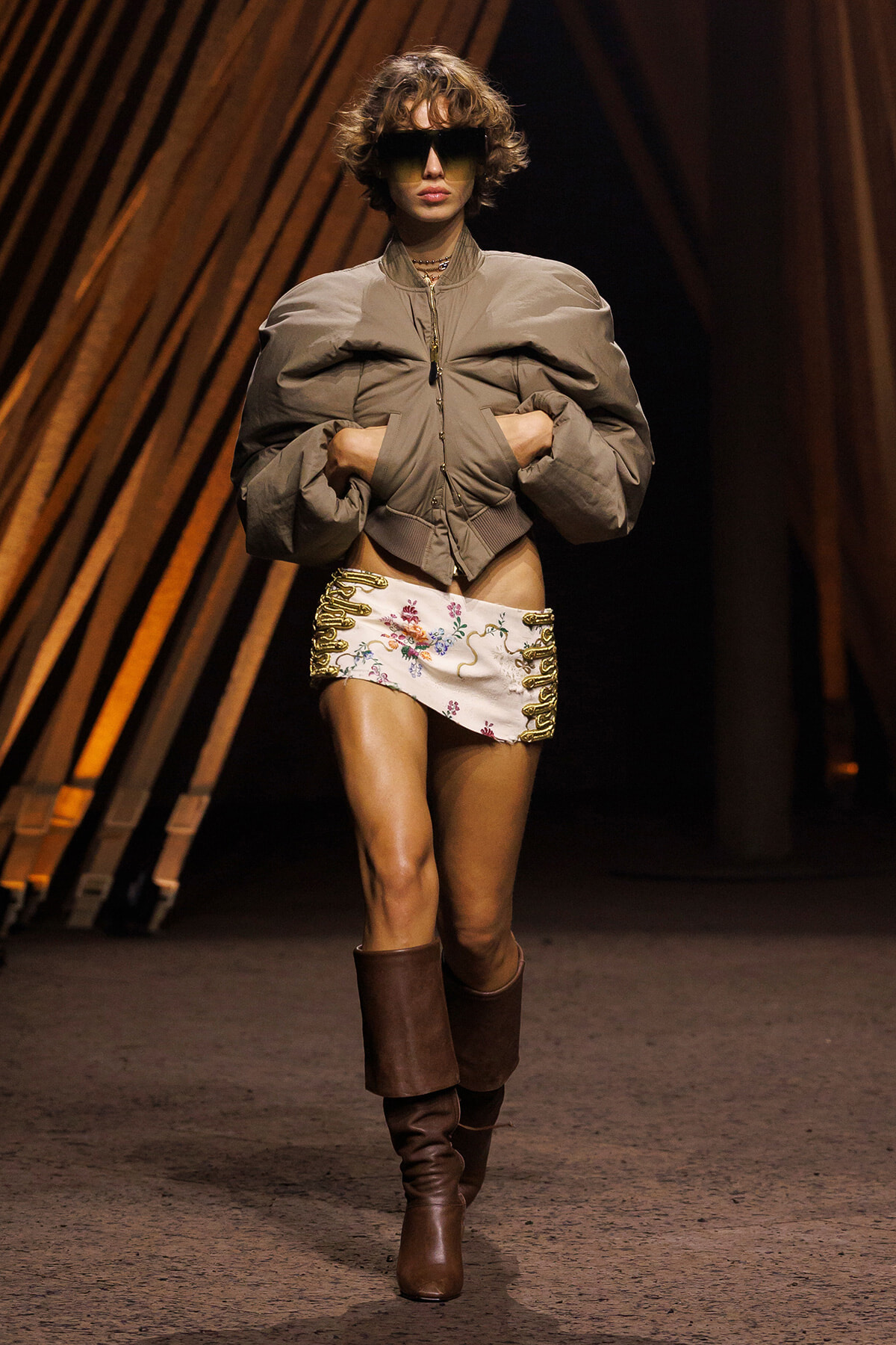 Model struts a runway in a taupe oversized jacket, a white embroidered mini skirt with gold lacing, and tall brown boots, wearing large sunglasses.