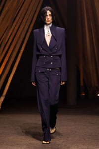 Runway model in a navy double‑breasted suit with structured shoulders, white lace neck top, and open‑toed sandals.