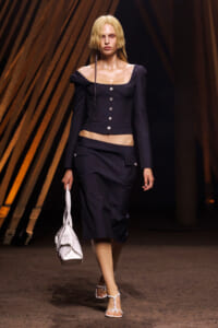 Model walking runway in a navy pinstripe two-piece outfit: off-shoulder cropped top with silver buttons and matching midi skirt, white handbag, and white strappy sandals.