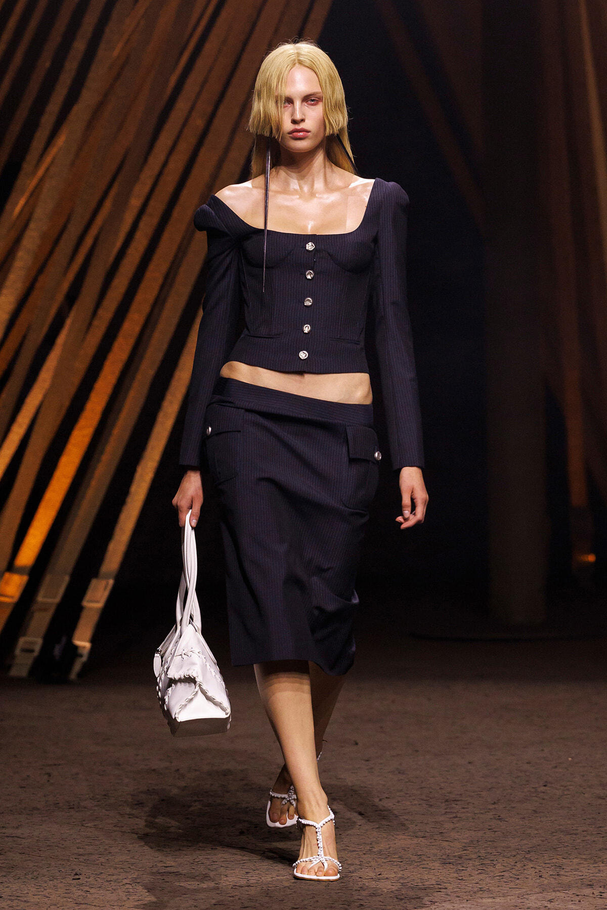Model walking runway in a navy pinstripe two-piece outfit: off-shoulder cropped top with silver buttons and matching midi skirt, white handbag, and white strappy sandals.