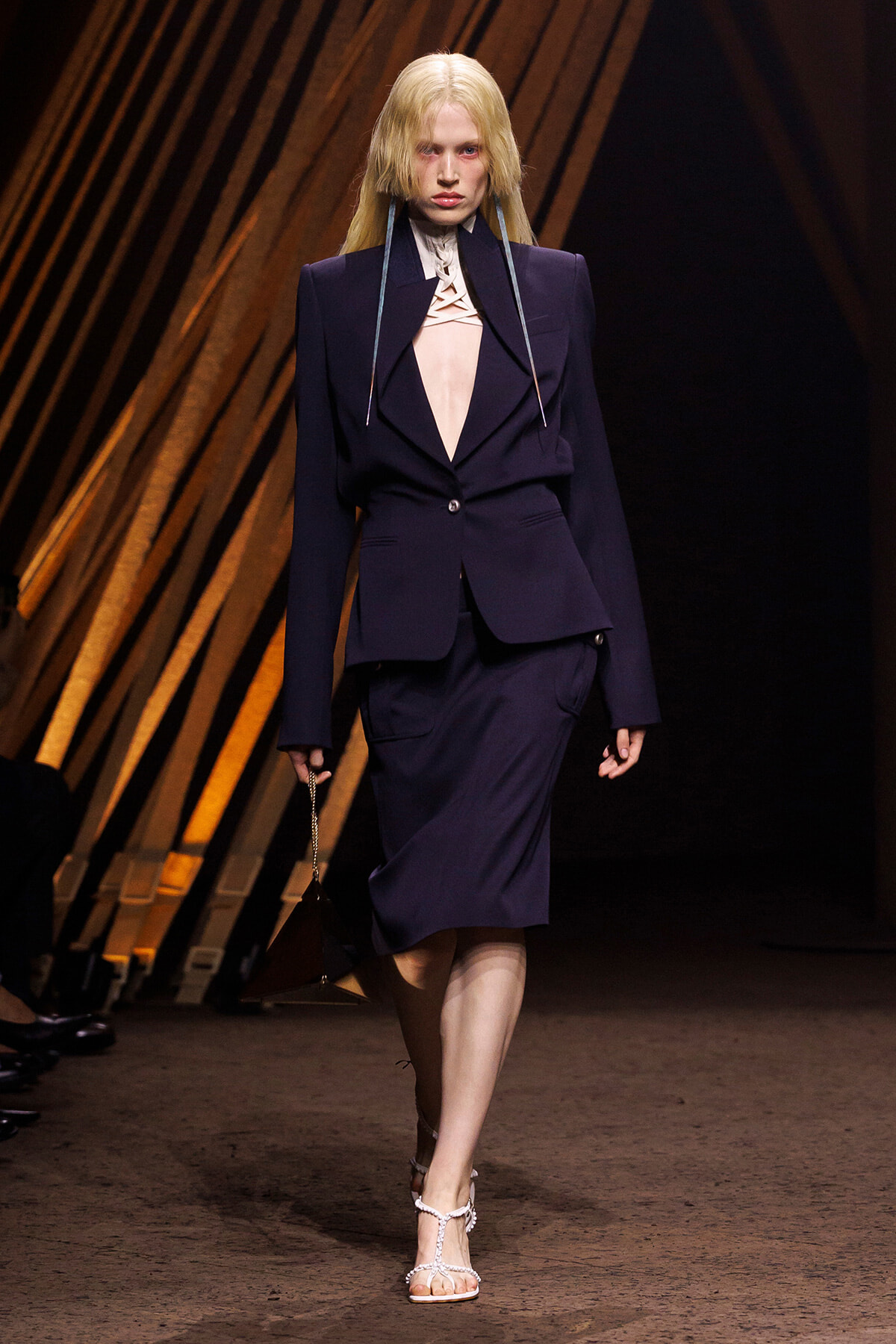 Model on a runway wearing a navy blazer and matching pencil skirt, with a cut-out blouse and long metallic ribbons around the neck, carrying a small triangular pendant bag and wearing white strappy sandals.