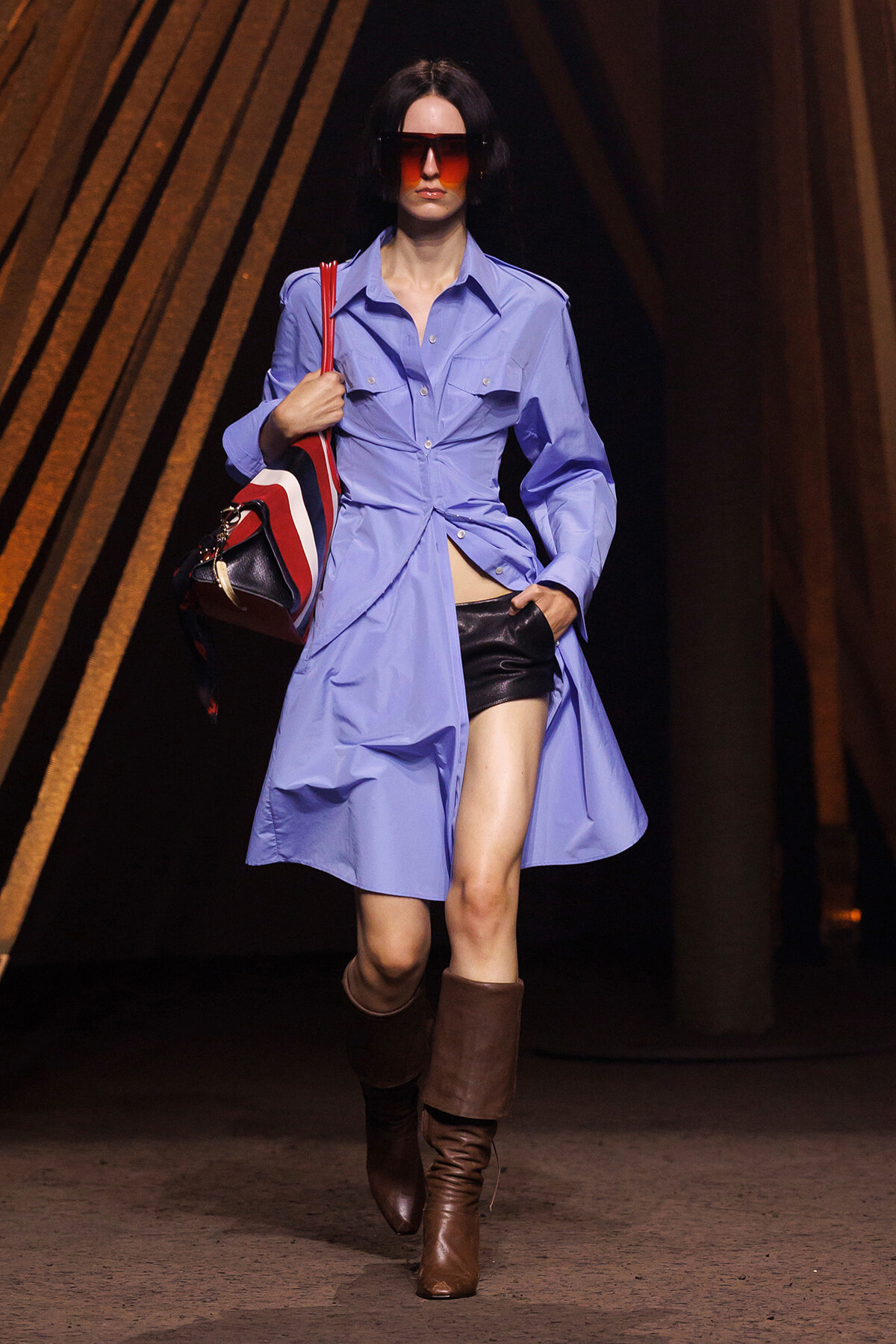 Model on a runway wearing a light blue shirt-dress with a knotted waist, black leather shorts, and brown knee-high boots, carrying a striped red–white bag and wearing red sunglasses.