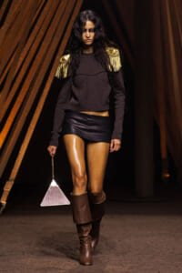 Model walking runway in a cropped dark sweater with gold fringe epaulettes, black mini skirt, and brown knee-high boots, carrying a triangular purse