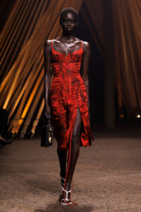 Model wearing a red satin dress with corset-style bodice and side slits walking a runway, holding a small black clutch.