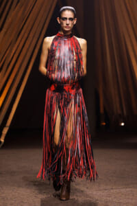 Model walks the runway in a red and black fringe dress with long vertical strips and boots.