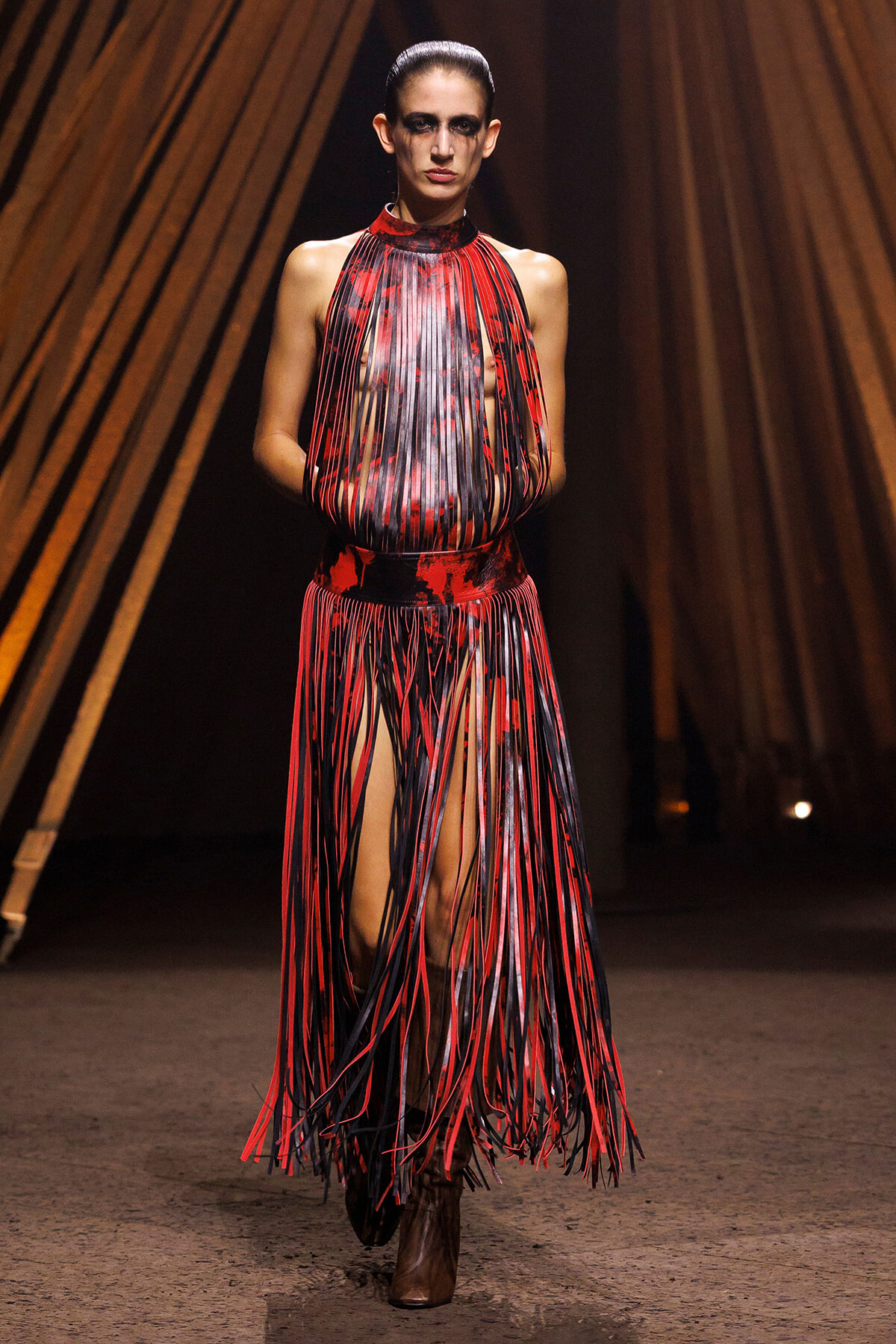 Model walks the runway in a red and black fringe dress with long vertical strips and boots.