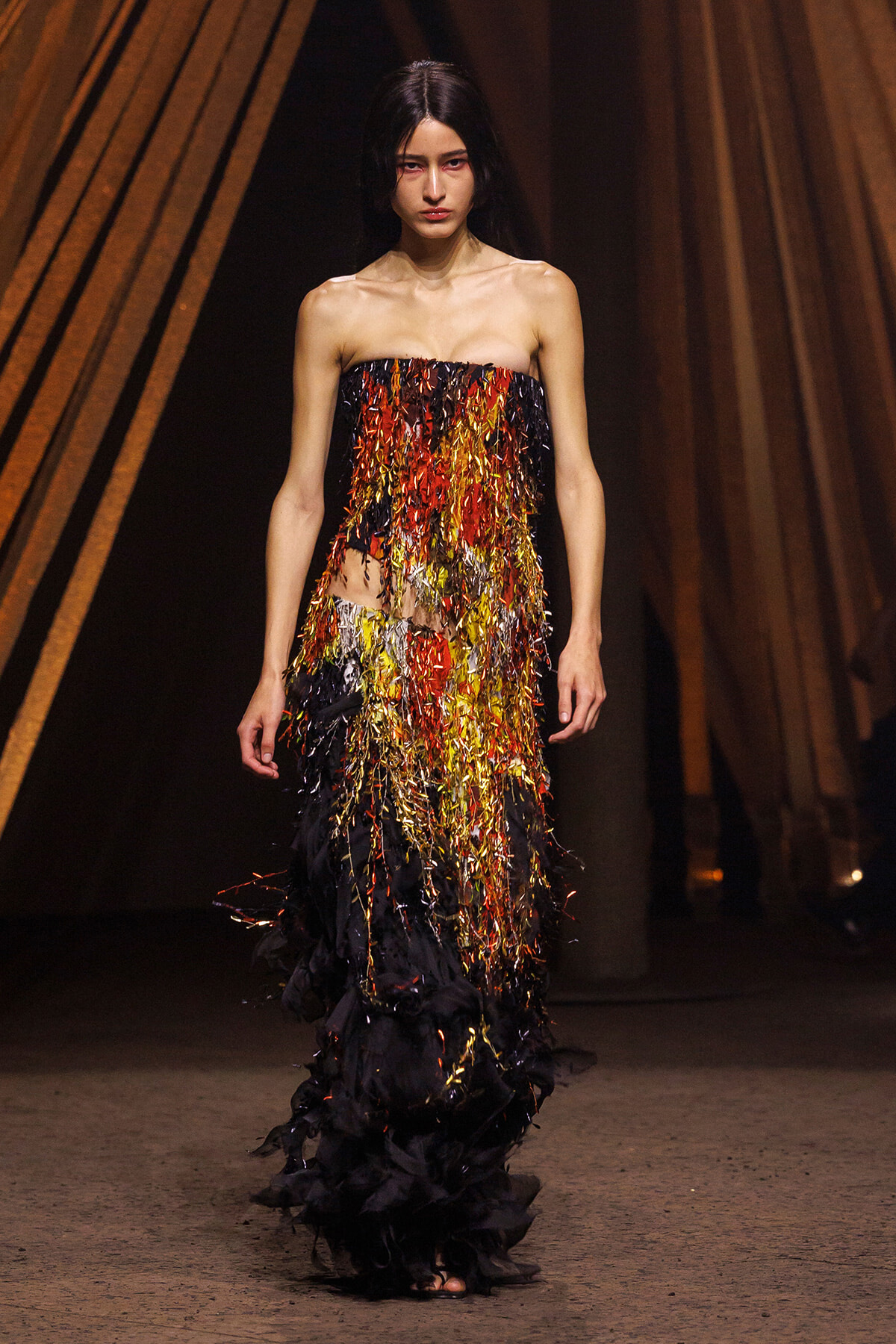 Model walks a runway in a strapless gown covered in multicolored fringe (red, gold, black) cascading to the floor against a dark backdrop.