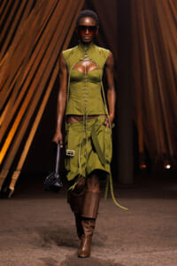 Model walking a runway in olive-green lace-up corset top with a matching cargo skirt, brown knee-high boots, oversized sunglasses, and a small black handbag.