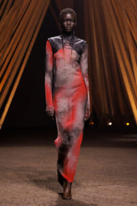 Model walks the runway in a long red and gray gradient knit dress with black leather shoulder detail on a dark stage backdrop.