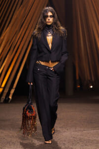 Model walks a runway wearing a black cropped blazer with a deep plunging neckline and matching high-waisted pants; she carries a long, fringed orange-black handbag and wears strappy sandals.