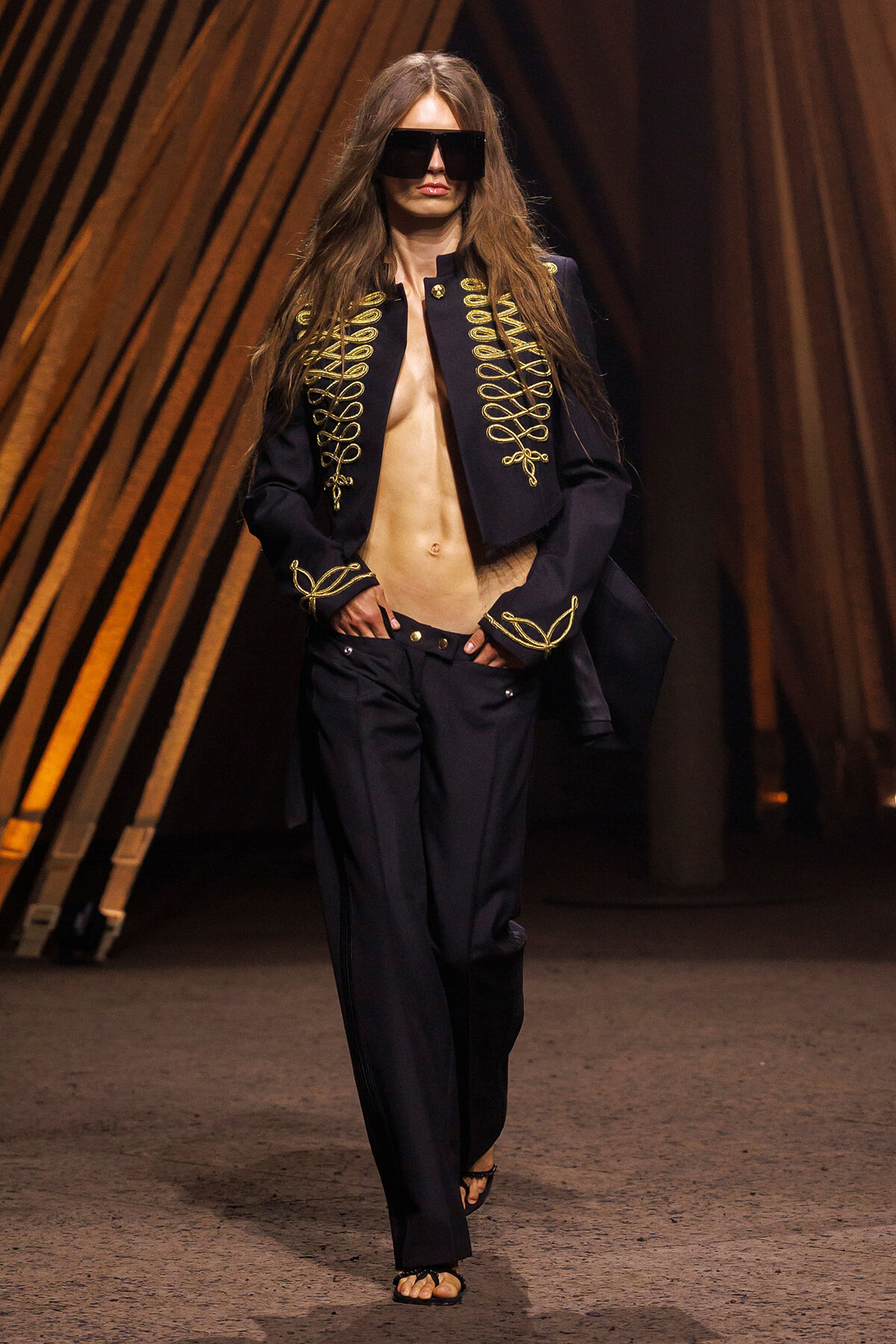 Model walking a fashion runway in a navy military-style jacket with gold braided trim, black wide-leg pants, and oversized black sunglasses; sandals visible as she poses mid-stride.