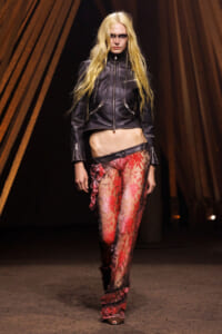 Model walks a runway wearing a cropped black leather jacket and sheer red lace pants with ruffles, blonde hair and dark makeup.