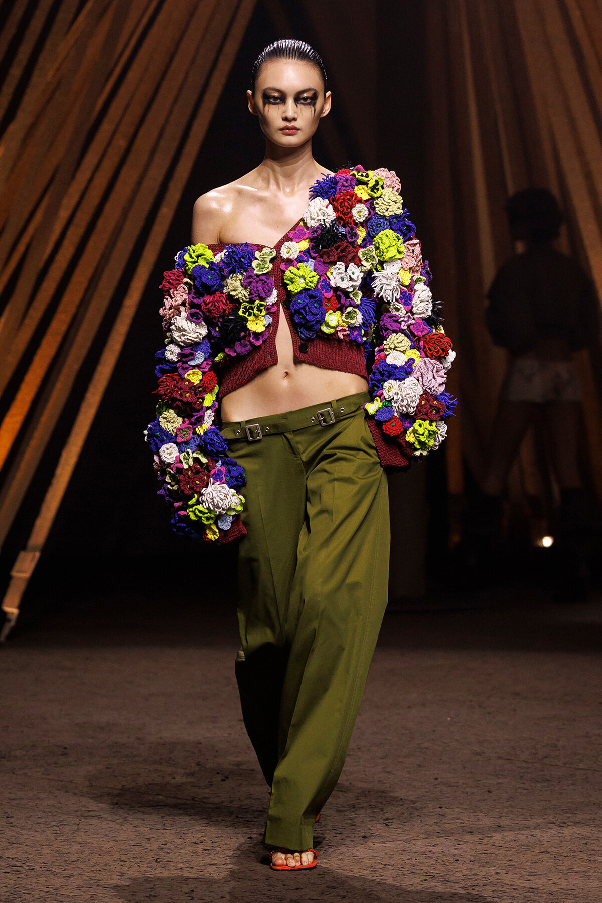 Model walks a runway in a bold, one-shoulder knit cardigan covered in multicolored yarn flowers, paired with olive-green wide-leg pants.