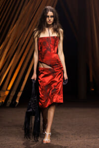 Model walking the runway in a red satin strapless dress with a subtle floral pattern, carrying a black fringed bag.