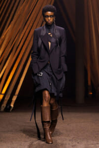 Model walks a fashion runway in an oversized black blazer, bandeau-style top, black cargo shorts with straps, and tall brown leather boots set against warm wooden backdrop.
