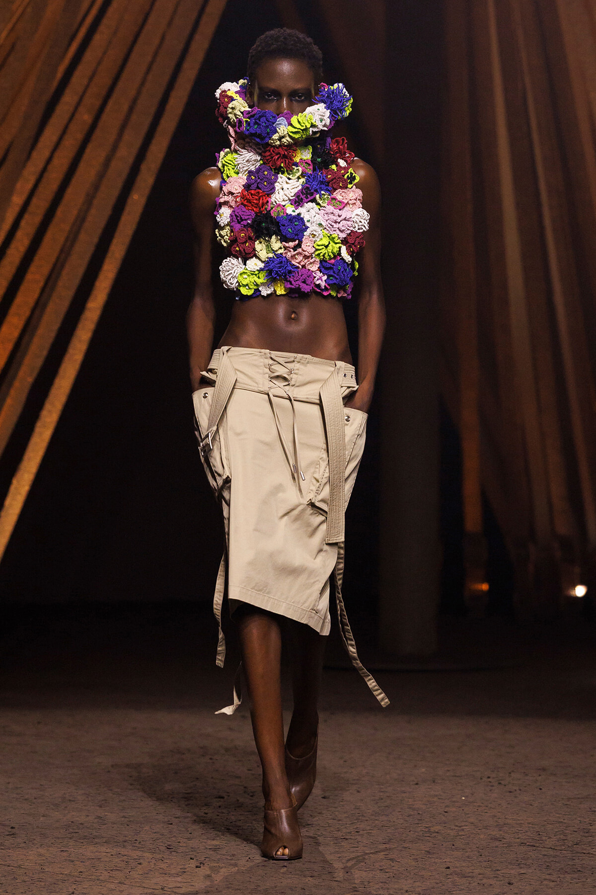 Model on a runway wearing a massive multicolored flower collar and chest piece with a beige utility skirt and peep heels.