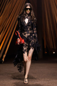 Model walking a runway in a sheer black lace dress, oversized sunglasses, and black strappy sandals, carrying a bright red handbag.