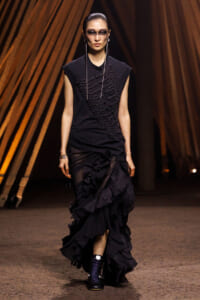 Model walking a runway in a black sleeveless top with ornate embroidery and a layered, ruffled black skirt; dark makeup and accessories visible.