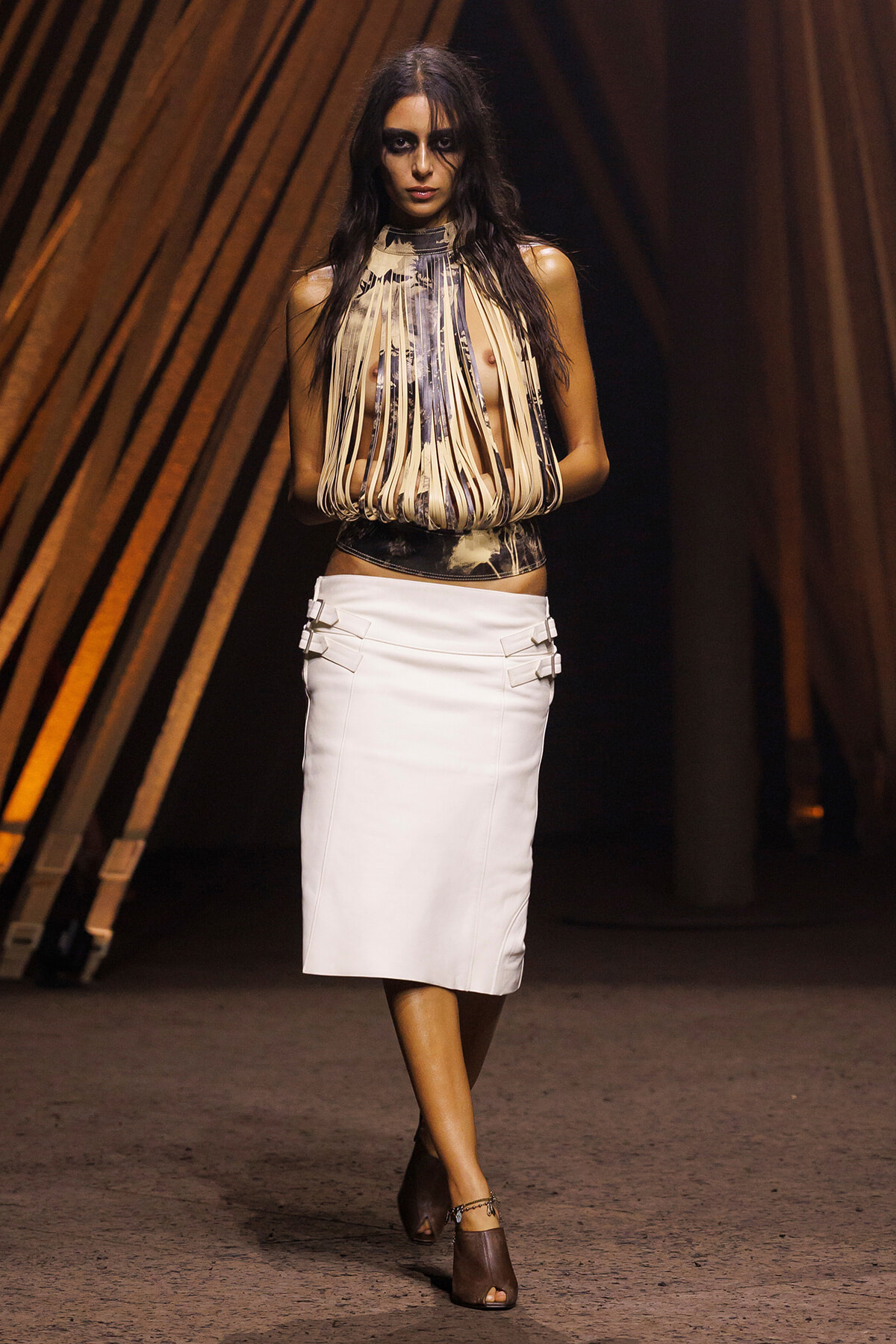 Model walks a fashion runway in a fringe-beige halter top and white pencil skirt with buckle details, brown open-toe heels, dark makeup.