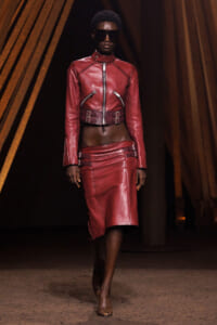 Model walks a fashion runway in a red leather two-piece set: cropped jacket with zippers and a matching knee-length skirt, wearing dark sunglasses.
