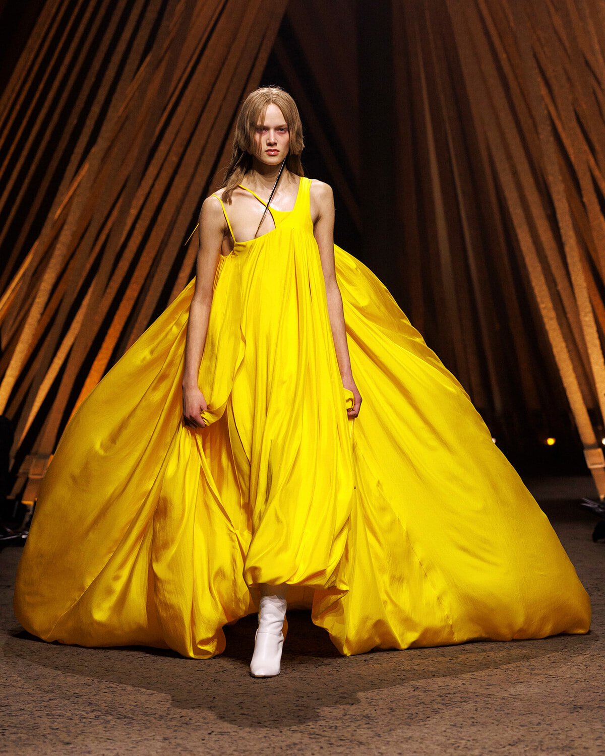 Model on a fashion runway wearing a vivid yellow, flowing, floor-length gown with a single-shoulder strap and white boots visible beneath the dress.