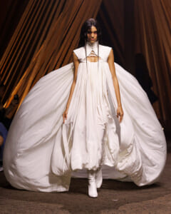 Model in a flowing white gown with a structured bodice and huge cape-like train on a runway with white boots.