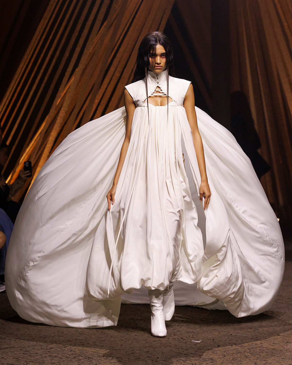 Model in a flowing white gown with a structured bodice and huge cape-like train on a runway with white boots.